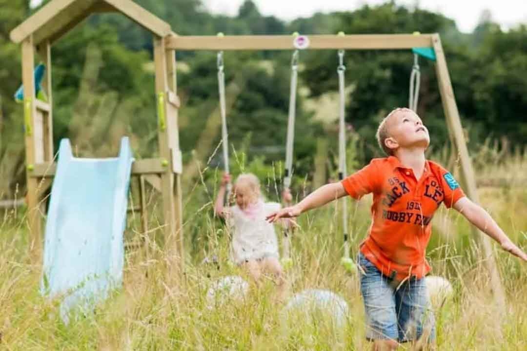 Children playing outdoors near a wooden swing set and slide in a natural garden setting from The Best Backyard Australia