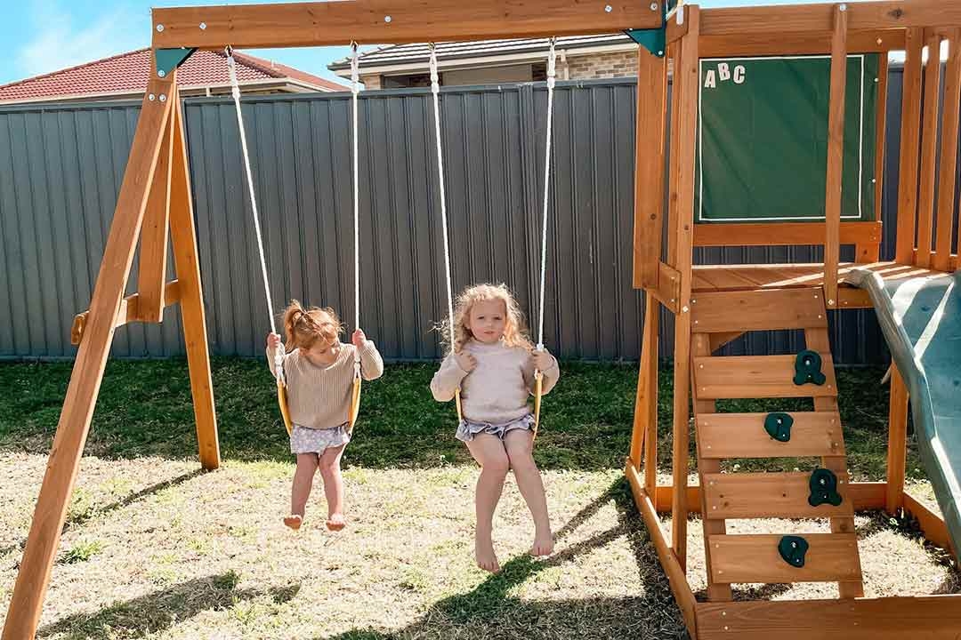 Two young girls swinging on a wooden backyard swing set with climbing wall purchased from The Best Backyard Australia