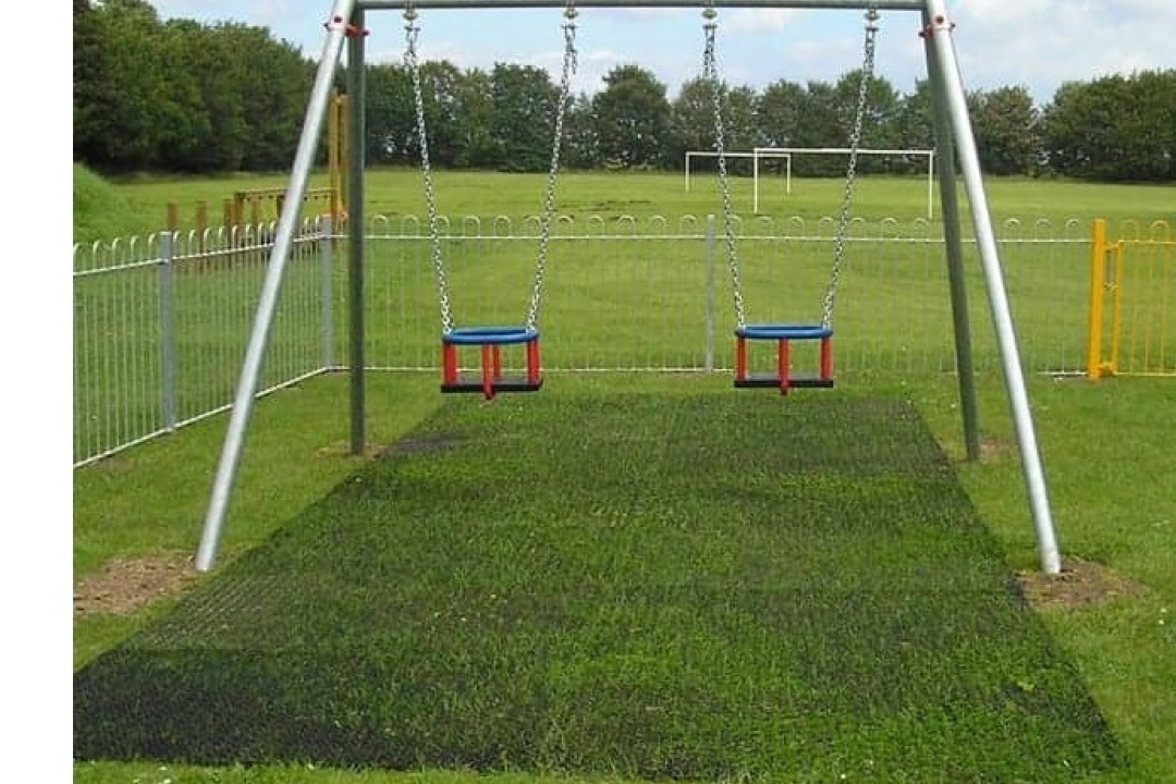 A rubber grass mat is installed beneath a double swing set on a grassy playground, providing a safe, natural-looking surface that protects the turf in the high-impact landing zone.