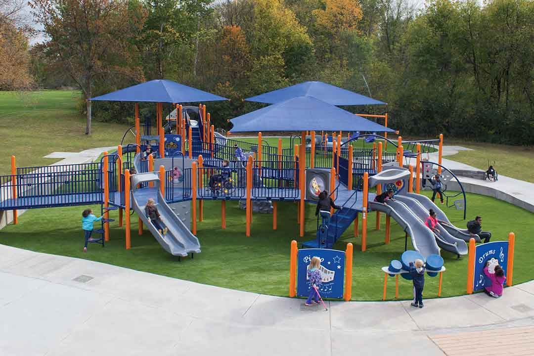 Inclusive accessible playground installed by St. Croix Recreation in Minnesota with children playing, multiple slides, shade structures, and outdoor musical instruments on artificial turf
