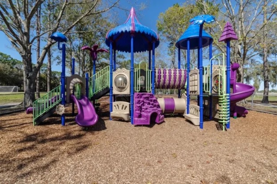  A whimsical purple and blue playground structure with slides, tunnels, and decorative towers, set on wood chip surfacing surrounded by tall trees in a park set…A whimsical purple and blue playground structure with slides, tunnels, and decorative towers, set on wood chip surfacing surrounded by tall trees in a park setting