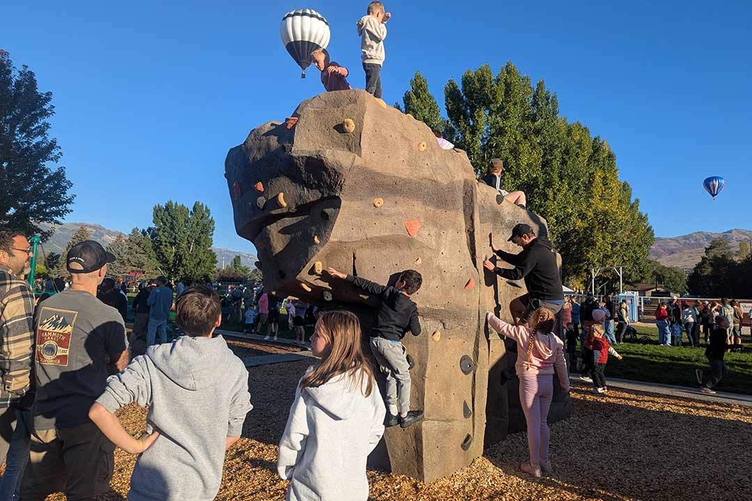 Families and children climbing a large EP Climbing Freeform community boulder at a park opening event with hot air balloons in the background