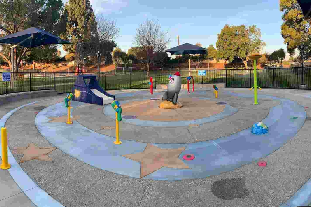 A space-themed splash pad featuring a central rocket sculpture, star-embedded flooring, and colorful water features set within a fenced circular park area.
