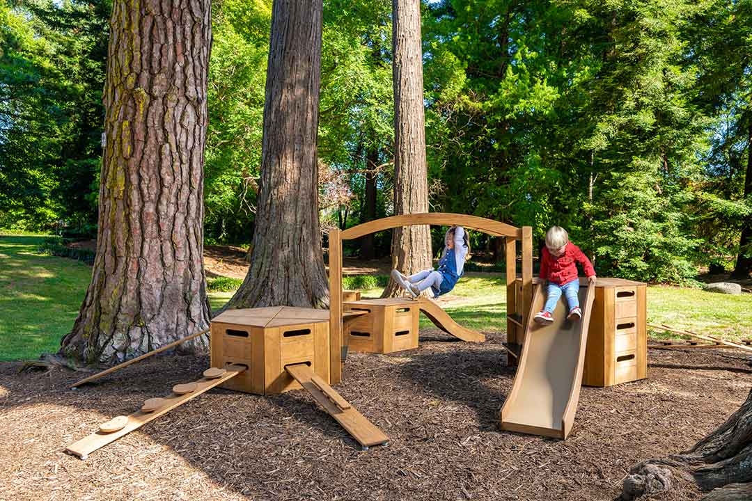 Two children playing on Outdorable modular timber play equipment with slide in a natural redwood forest setting