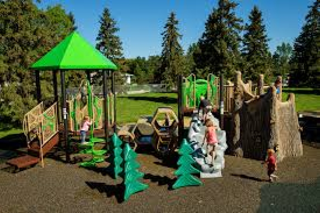 A nature-themed playground features wooden structures, green tree sculptures, and climbing elements nestled among mature trees in a lush park setting.