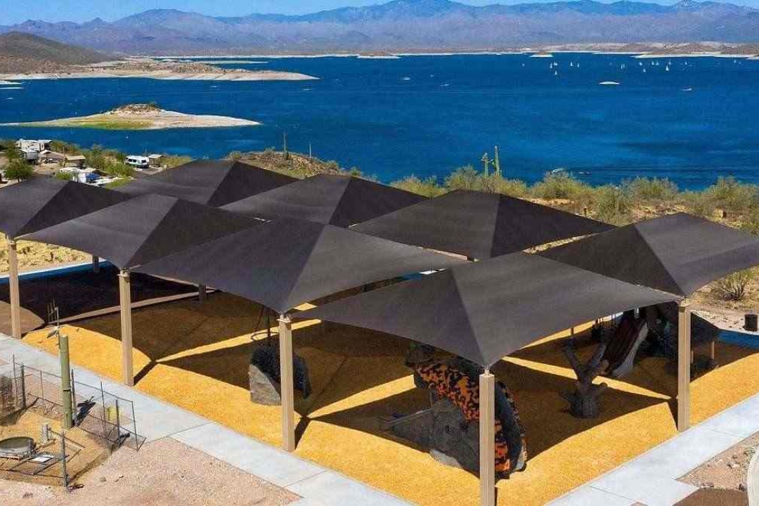 A stunning geometric shade structure covers a playground area with golden rubber surfacing, dramatically set against a sweeping desert lake backdrop.