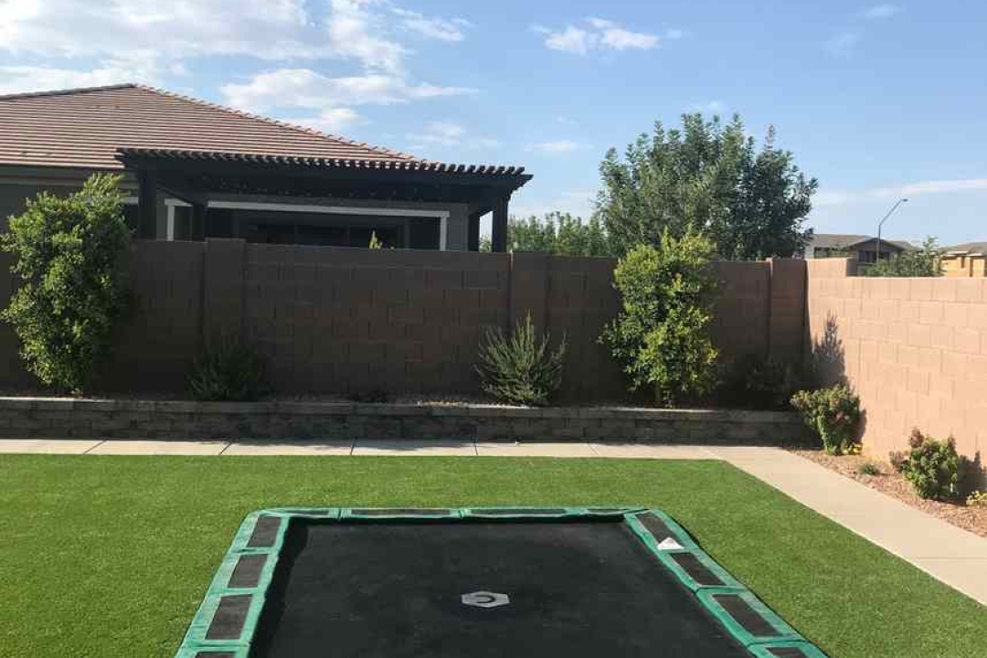 A Capital Play rectangular in-ground trampoline installed flush with a lush green artificial grass backyard in Texas. The trampoline features green safety padding around the edges and blends seamlessly into the outdoor space, with a beautiful landscaped yard, stone retaining wall, and pergola visible in the background.