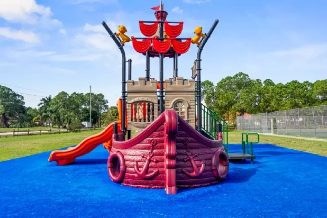  A pirate ship-themed playground structure with red and brown details, an orange slide, and anchor decorations, set on bright blue rubber safety surfacing in a …A pirate ship-themed playground structure with red and brown details, an orange slide, and anchor decorations, set on bright blue rubber safety surfacing in a park.