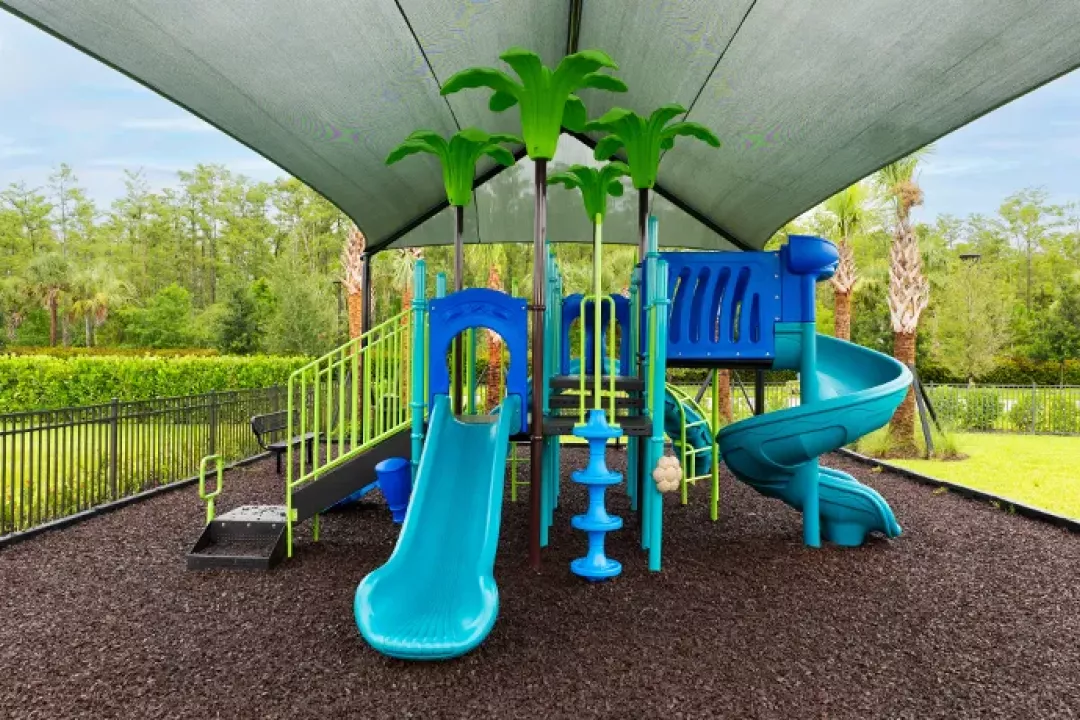 A vibrant blue and teal playground with slides, climbing features, and decorative palm tree toppers, sheltered under a large shade canopy and set on dark rubber mulch surfacing surrounded by lush greenery.