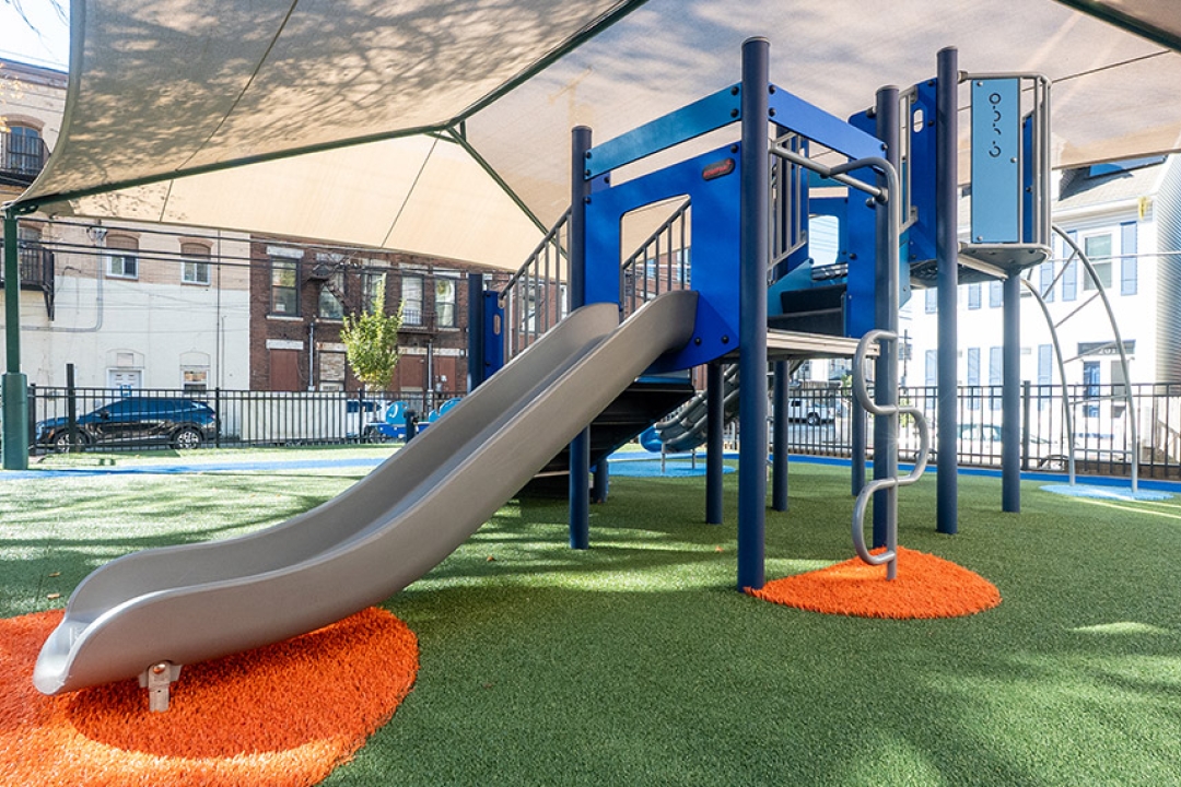 A vibrant blue commercial playground structure at the Watson Institute in Sharpsburg, PA, featuring a stainless steel slide, interactive panels, and colorful orange and green artificial turf surfacing, all sheltered beneath a large shade sail structure. This thoughtfully designed urban play space reflects YOCCA Playgrounds + Outdoor Fitness's dedication to creating safe, engaging, and visually striking environments for children in the communities they serve.