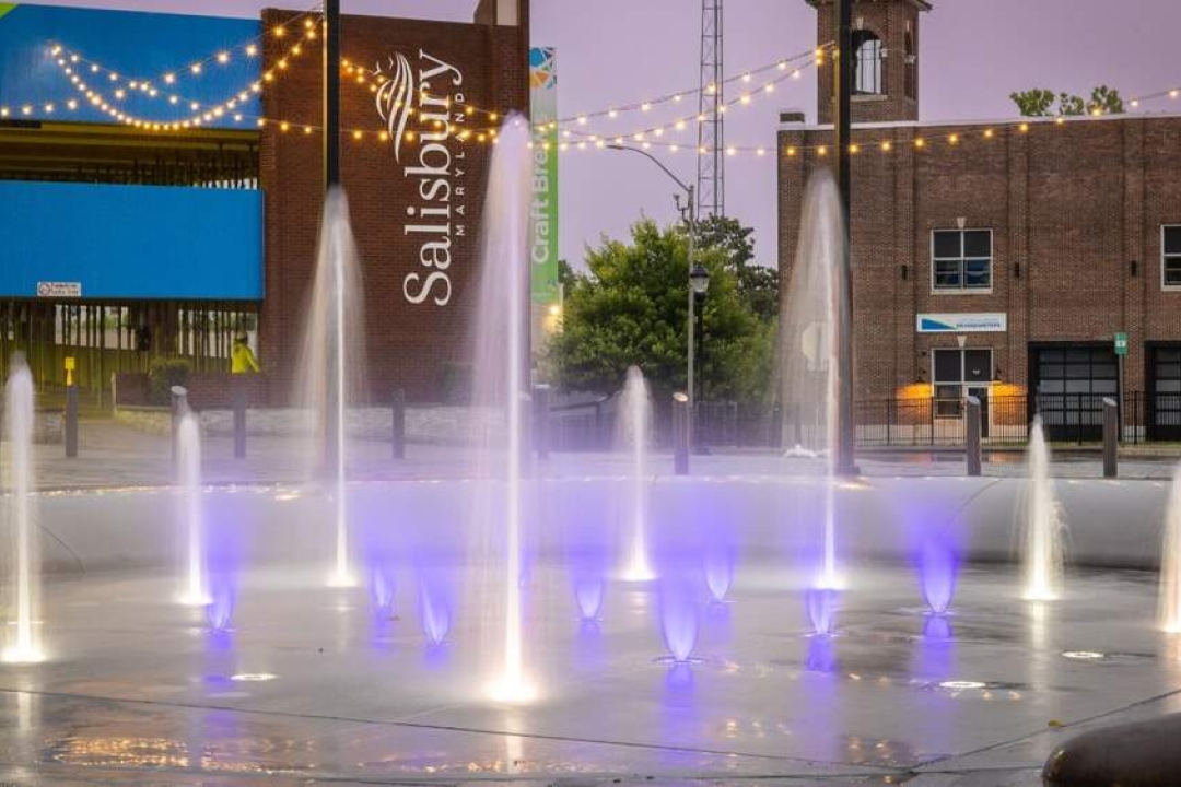 A vibrant purple-lit splash pad with dancing water jets lights up the evening in downtown Salisbury, Maryland.