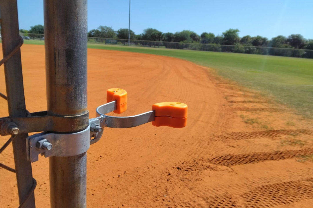 A KinderLatch safety gate latch with bright orange polymer covers installed on a chain-link fence at a baseball field, demonstrating its flush, low-profile design in a real-world sports setting.
