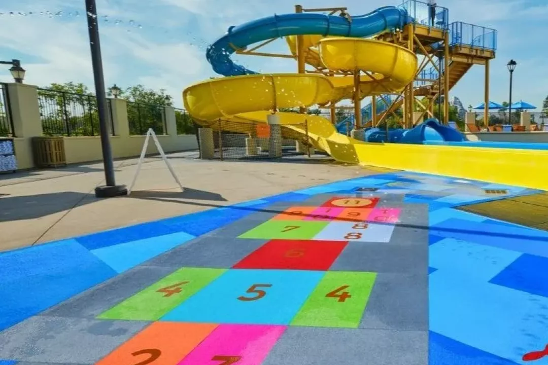 A vibrant water park featuring a colorful numbered hopscotch safety surface flooring in the foreground with a bold yellow and blue multi-lane water slide struc…A vibrant water park featuring a colorful numbered hopscotch safety surface flooring in the foreground with a bold yellow and blue multi-lane water slide structure in the background.