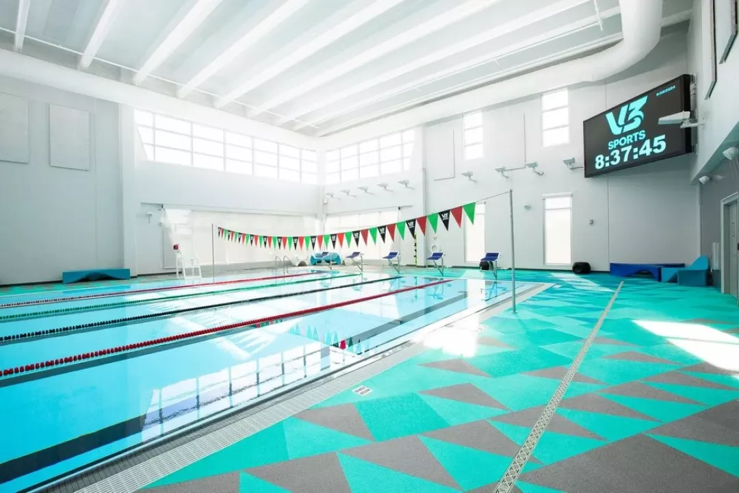 A modern indoor competitive swim facility featuring Life Floor's vibrant teal and gray geometric safety surfacing along the pool deck, with lane dividers, team banners, and a V3 Sports scoreboard overhead.