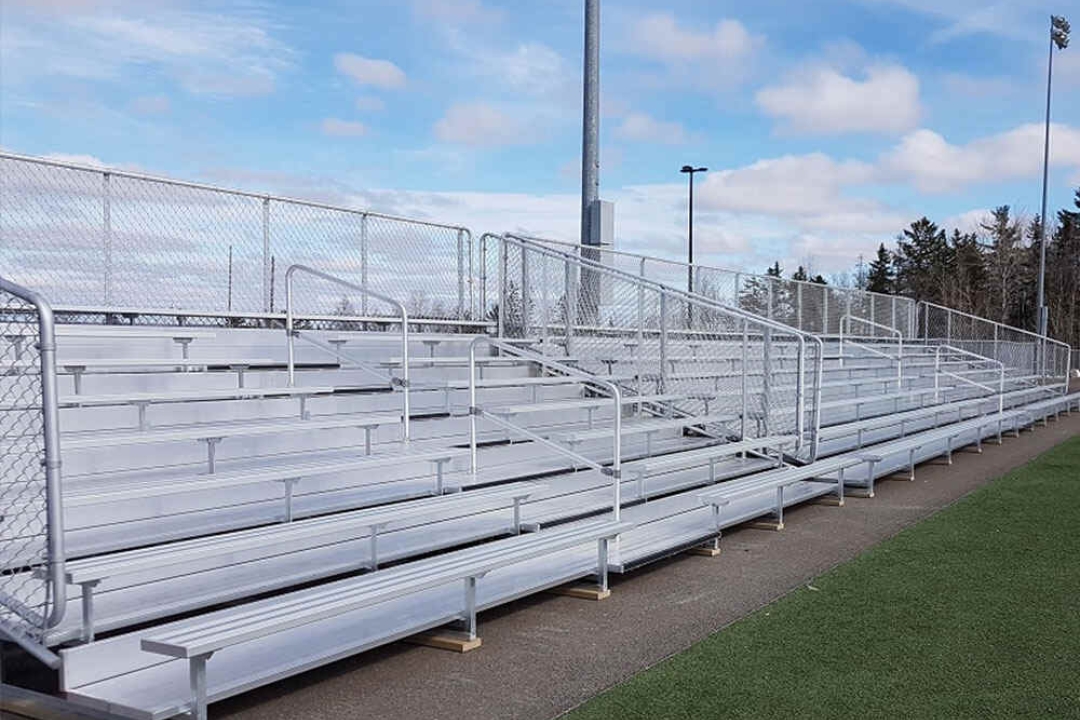 Multi-tiered aluminum bleacher seating with safety railings lines the edge of an outdoor sports field under a partly cloudy sky.