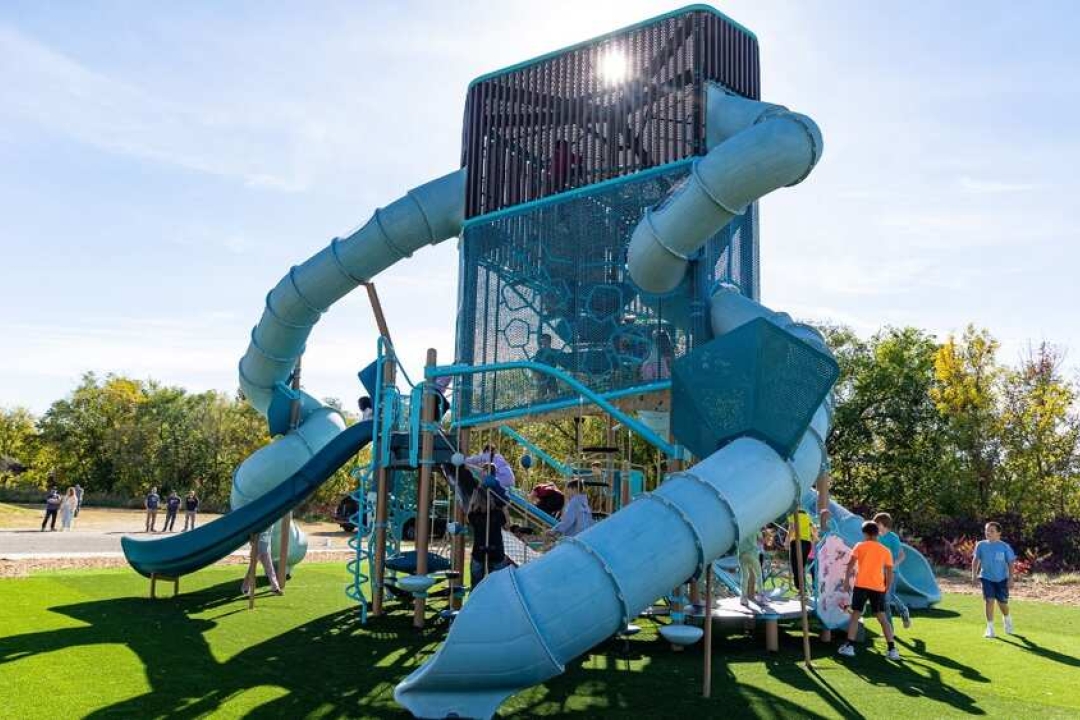 Children enjoy a towering teal playground structure featuring multiple tube slides, climbing nets, and open play areas on a sunny day.
