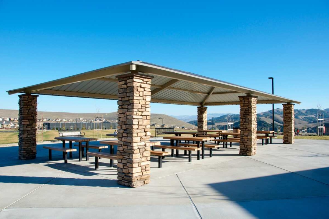 A spacious open-air park pavilion with stacked stone columns and a metal roof provides shaded picnic seating with sweeping mountain views in the background.