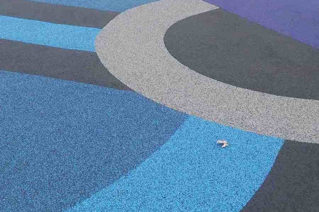A vibrant multi-colored poured rubber playground surface features bold swirling patterns in blue, purple, grey, and yellow.