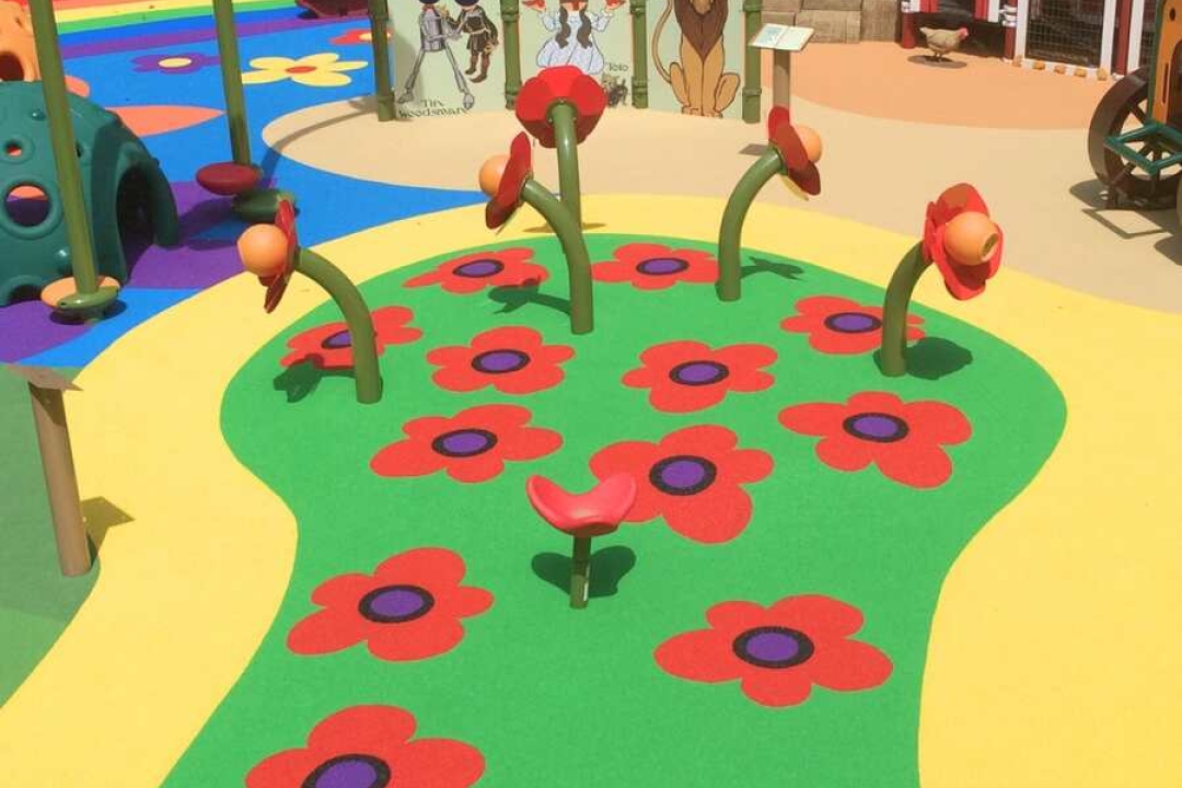 A whimsical Wizard of Oz-themed playground showcases a poppy field rubber surface design with red flowers on a winding yellow and green path.