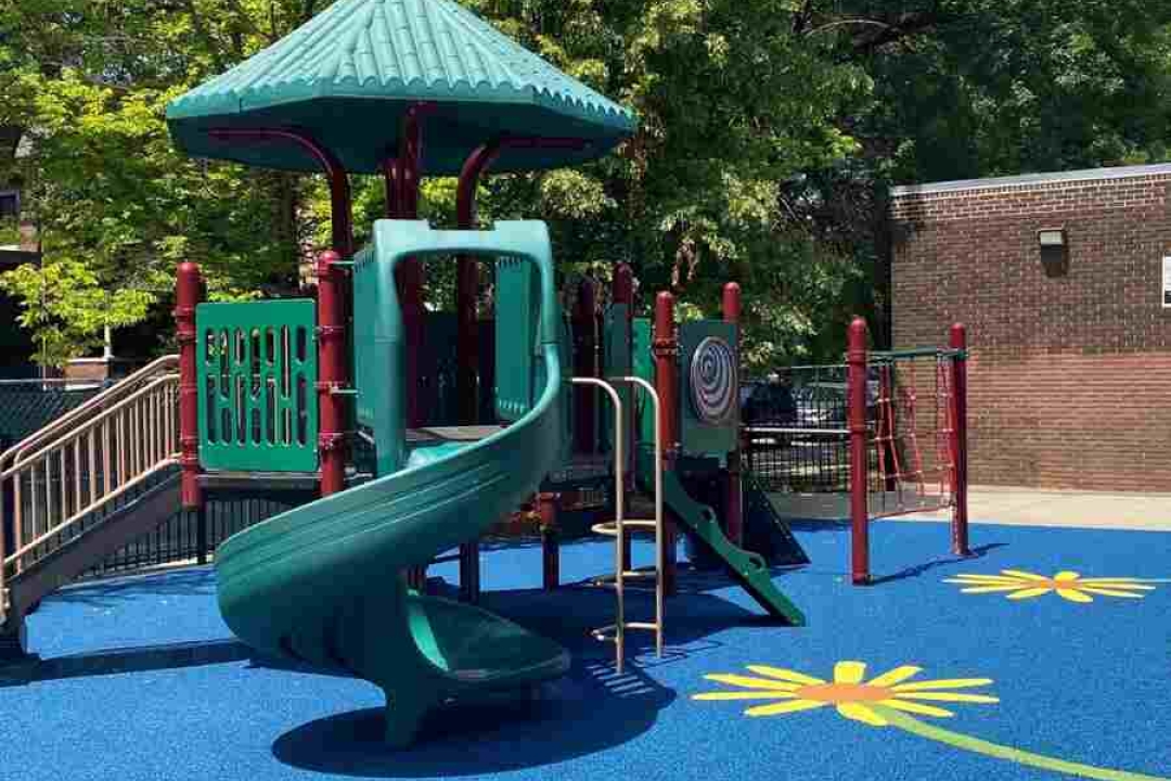 A teal and red playground structure with a spiral slide sits on a bright blue rubber surface decorated with cheerful yellow flower designs