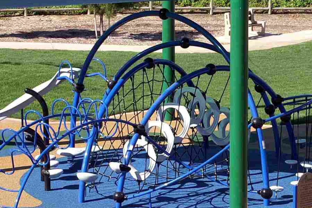 A creative blue arched net climbing structure with spiral handholds is shaded by a navy sail canopy over colorful poured rubber surfacing.
