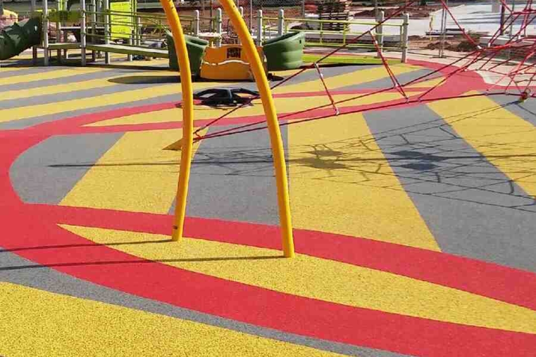 A bold yellow and red striped rubber playground surface pairs with a bright yellow arch climber and red rope net during an exciting new park installation.