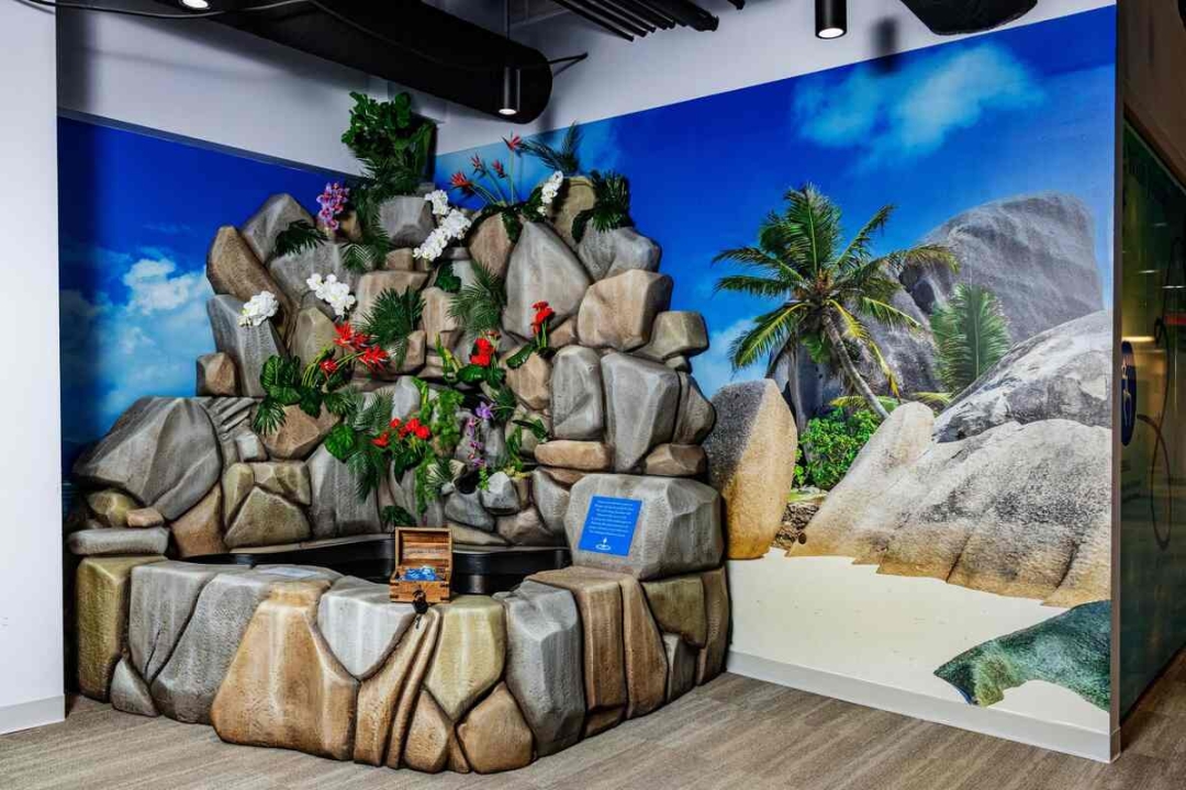 A stunning 3D sculpted rock formation with tropical flowers and foliage sits against a photorealistic beach mural backdrop.