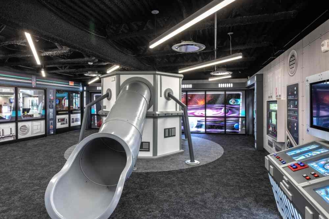 A fully immersive space station-themed room features a metal tube slide, mission control panels, and galaxy murals for an out-of-this-world experience.