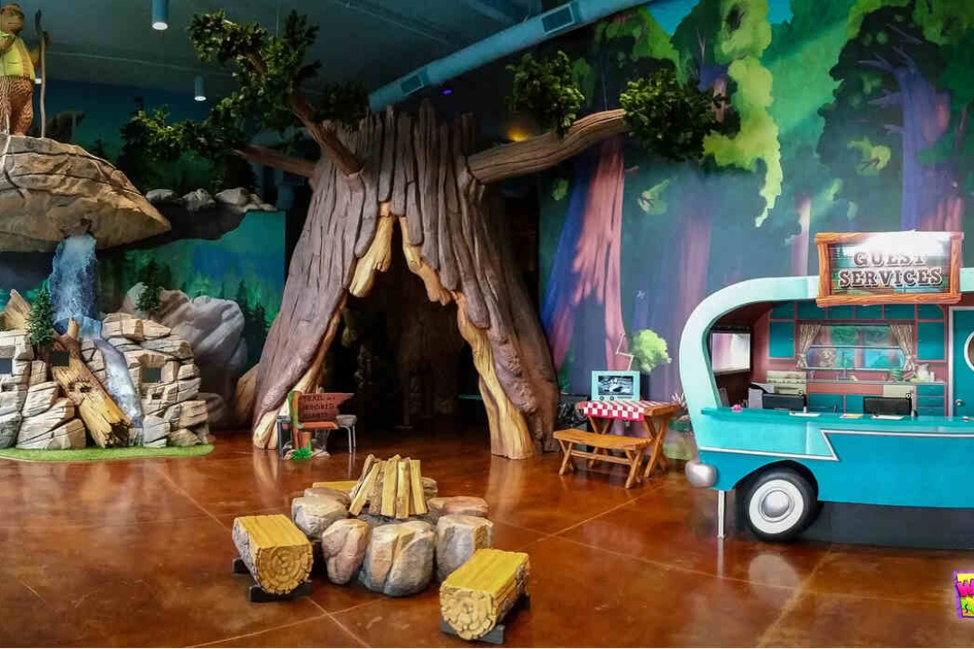 A breathtaking indoor camping-themed environment features a massive sculpted tree, rock formations, a vintage camper guest services desk, and a sweeping forest mural.