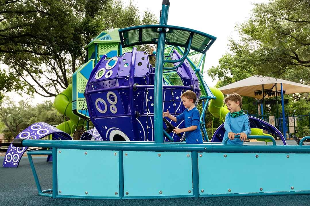 Two boys playing on the GameTime ship-themed interactive play panel at Carol Fox Park Jersey Village Texas with the kraken climber in the background