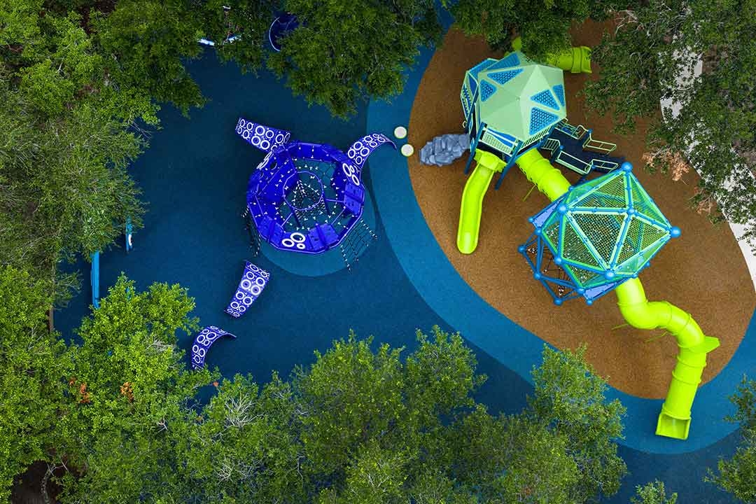 Aerial view of the complete Carol Fox Park playground layout showing the GameTime kraken climber and multi-level play tower with tube slides surrounded by oak trees in Jersey Village Texas