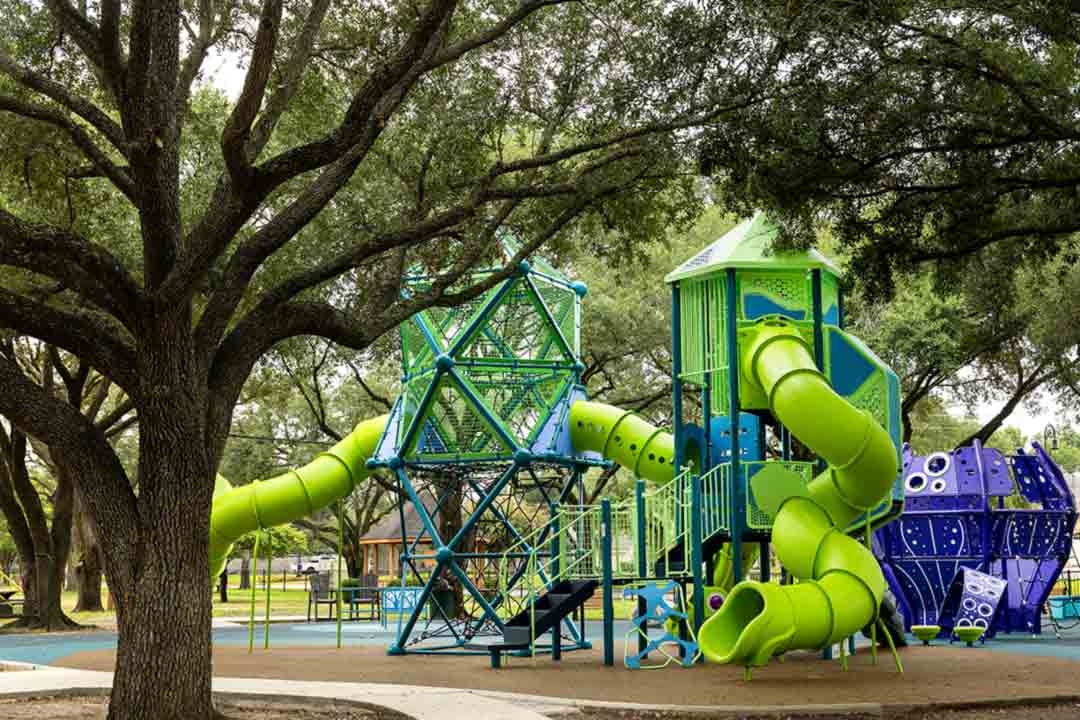 GameTime multi-level play tower with lime green tube slides and net climber at Carol Fox Park Jersey Village Texas under a canopy of mature oak trees