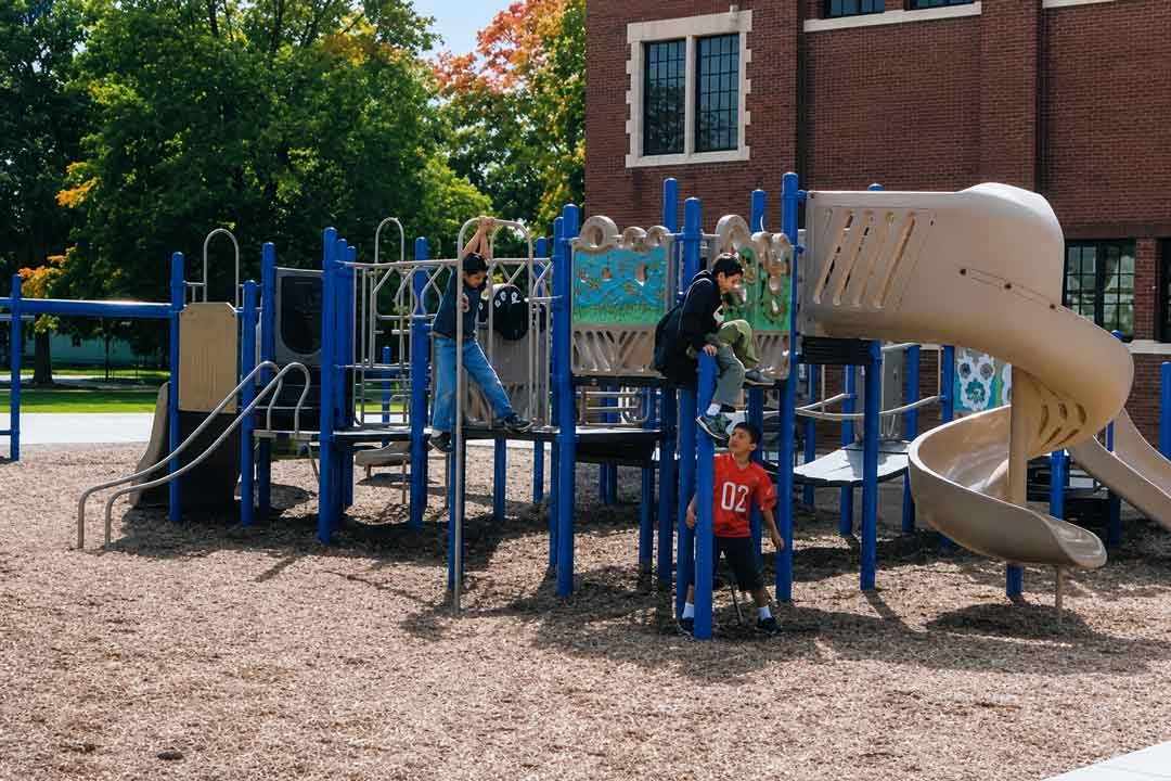 Children playing on a modern blue-and-tan playground structure outside a brick school building on a sunny day, with slides, climbing features, and wood chip surfacing surrounded by green trees.